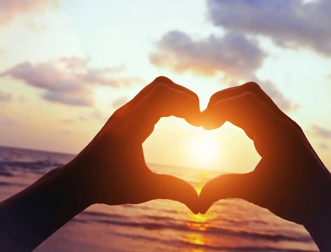 Hands forming a heart shape framing the sunset over the ocean.